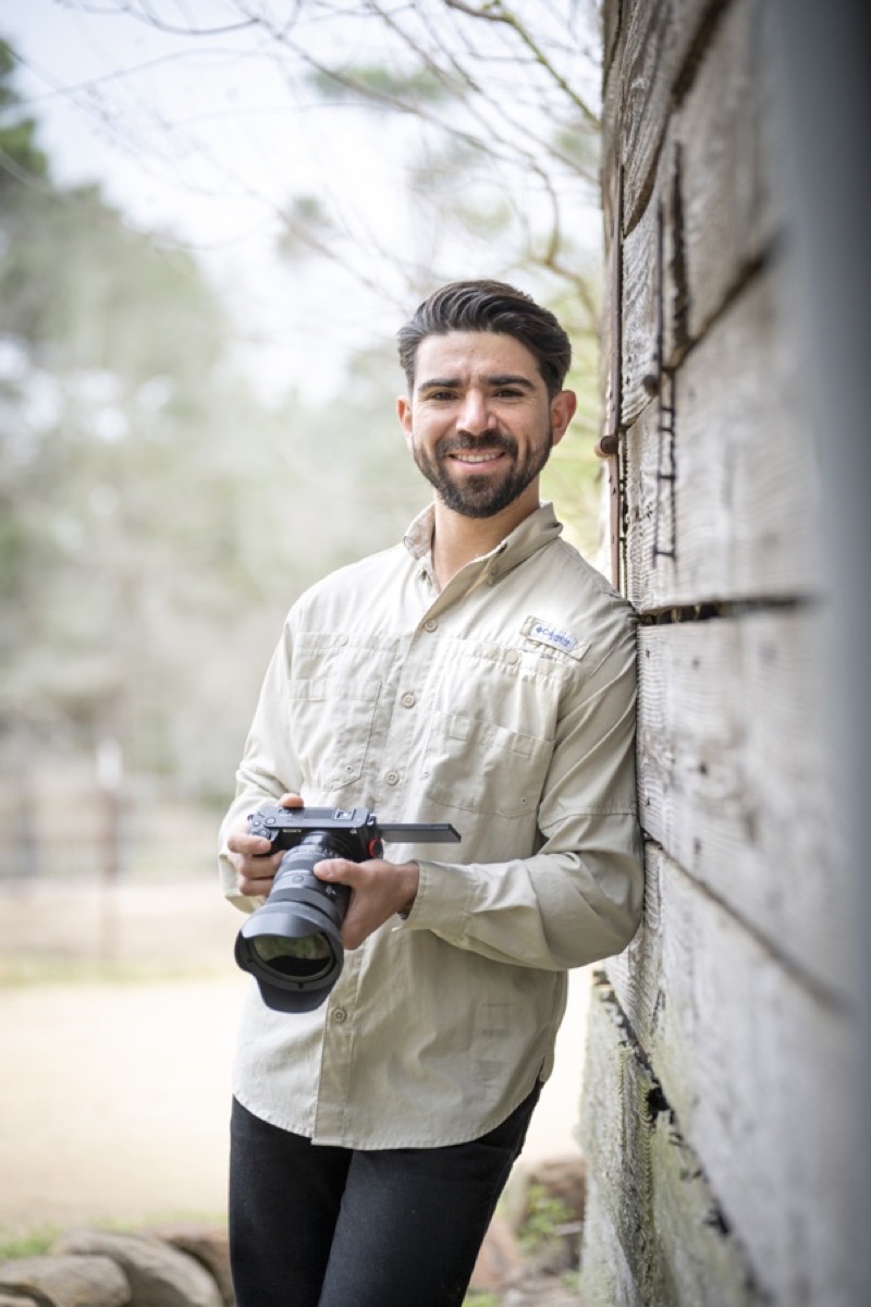 Aaron Amezcua standing outdoors with a camera in hand, beside a weathered wooden barn wall.