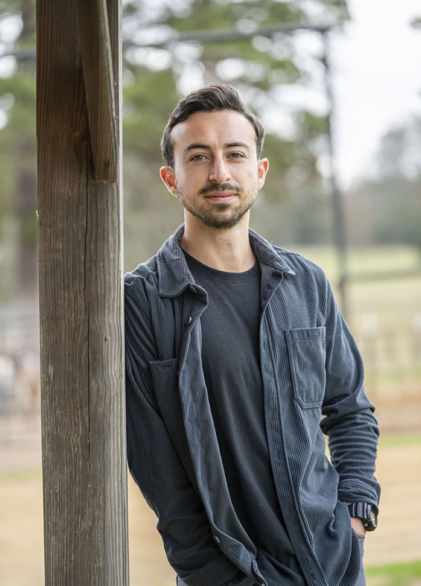Erick Amezcua leaning against a wooden post on the farm, pasture and pine trees behind him.